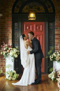 Bride and groom sharing a kiss in front of a red door with floral arrangements.
