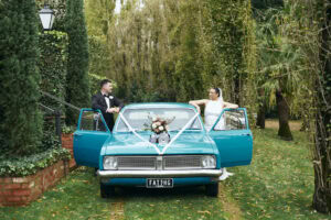 Bride and groom with a vintage blue car in a lush garden setting.