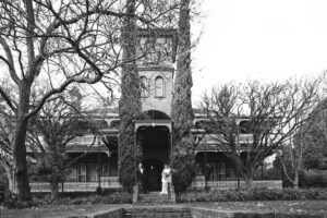 Historic Victorian-style house with large trees and a couple in wedding attire.