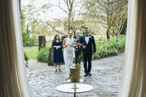 Wedding couple arriving at outdoor venue with guests and lush garden backdrop.